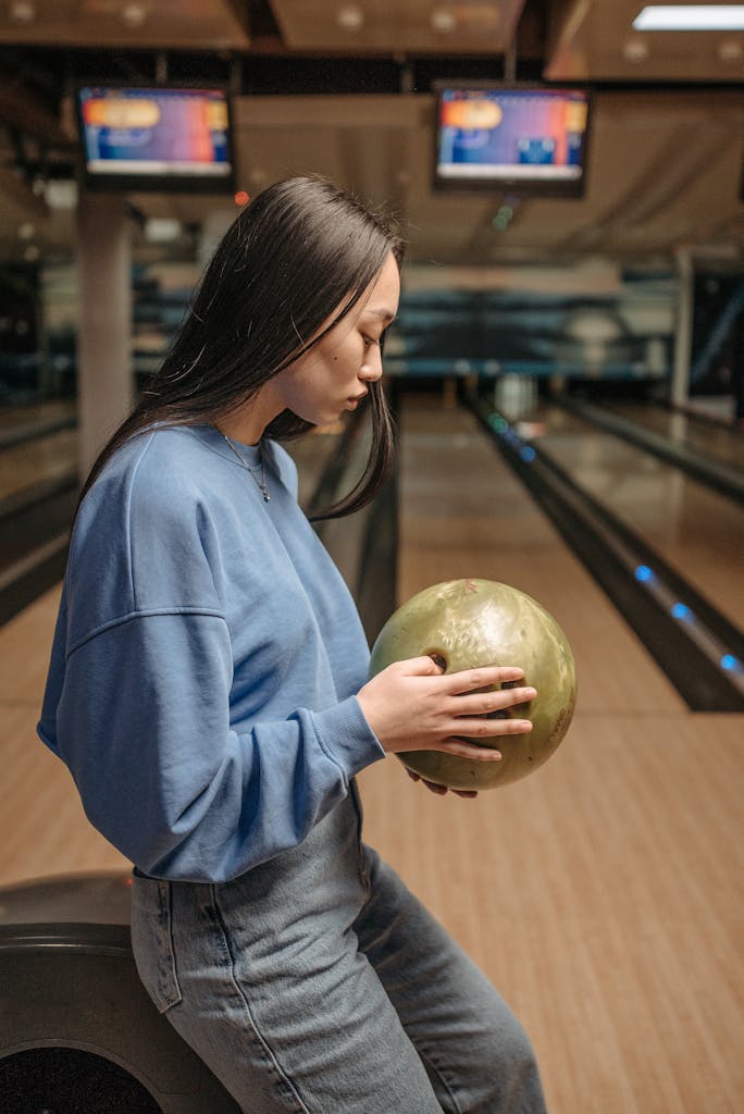A woman in a blue sweater holds a bowling ball at an indoor bowling alley, focusing on her grip.
